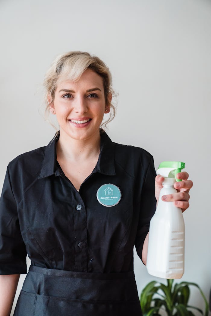 services-03 Smiling woman in uniform holding a spray bottle, symbolizing professional cleaning services indoors.